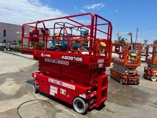 A red LGMG AS0812E scissor lift parked outdoors on a paved lot, with other similar lifts visible in the background.
