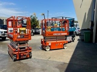 Two orange scissor lifts parked on an outdoor concrete lot next to a building.