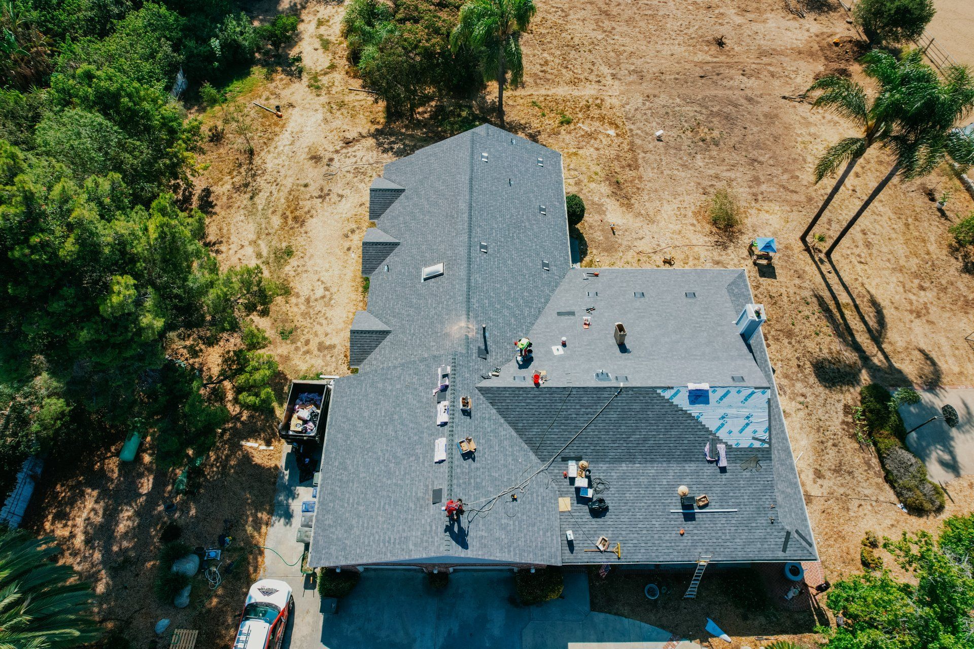 Aerial view of roof workers on a gray house surrounded by trees and dry ground