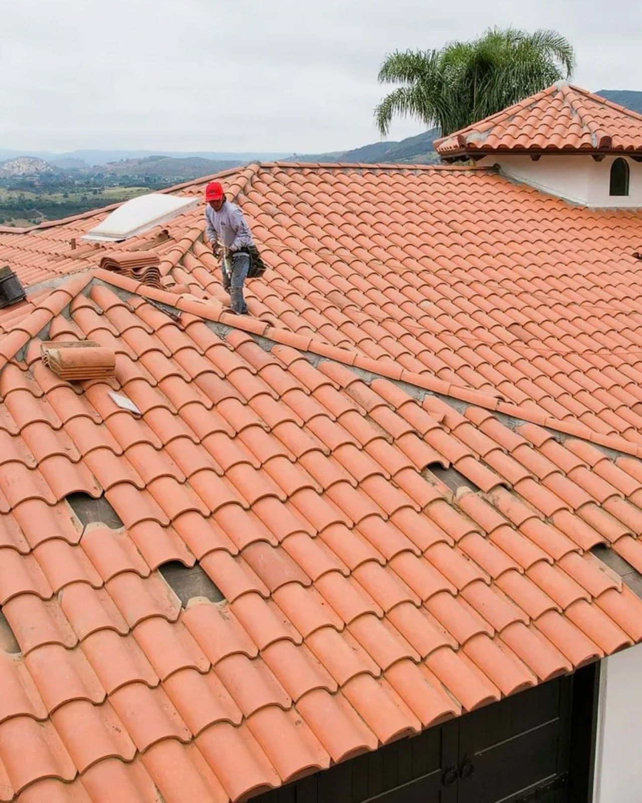 Worker on a red tile roof, inspecting or repairing near a chimney with a hillside view in the background