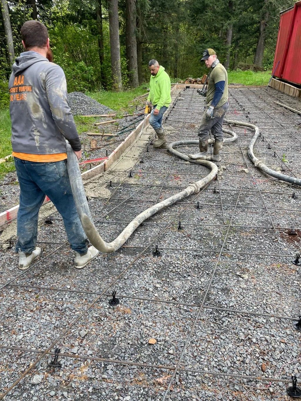 Three workers use a concrete pump hose to pour foundation material over a gravel site with evenly spaced stakes.