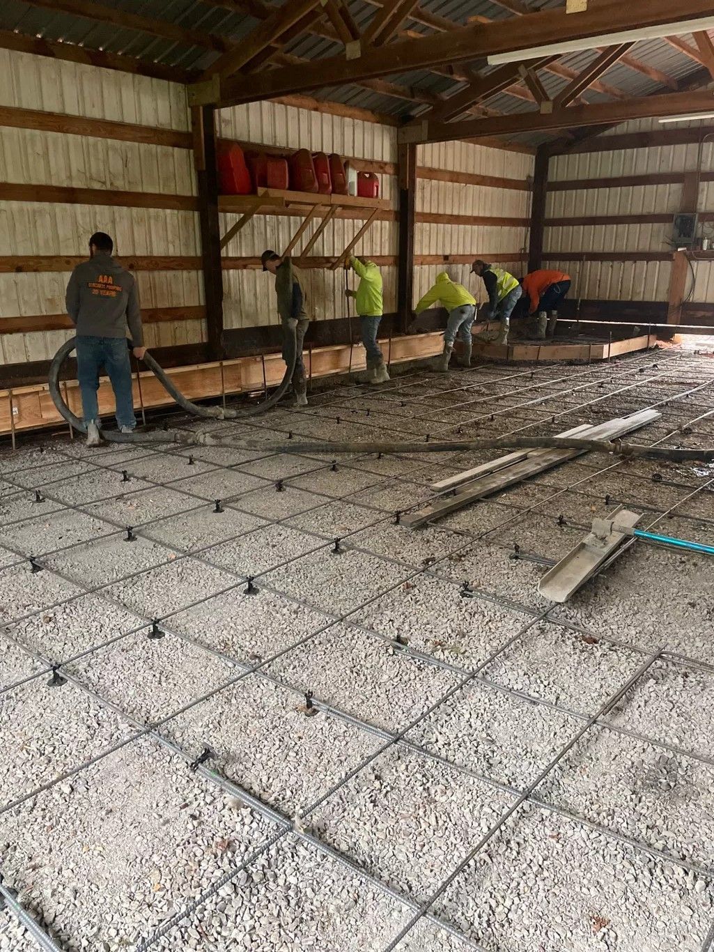 Workers in a large, insulated shed prepare a concrete foundation, laying rebar grid over a gravel sub-base.