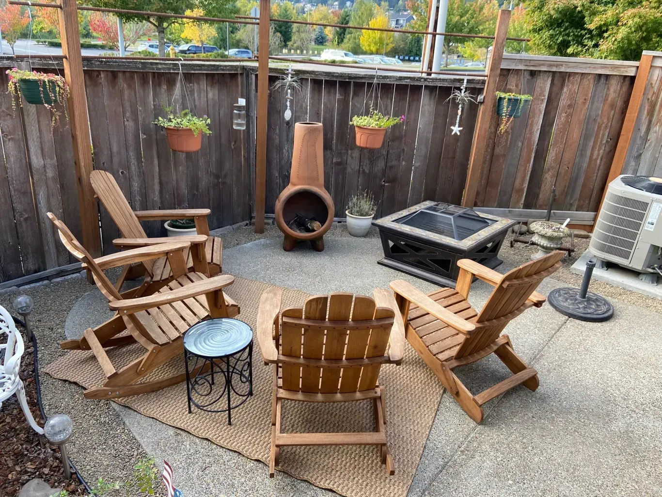 A patio setup with three wooden rocking chairs, a small metal table, a fire pit, and a clay chiminea on a woven rug.