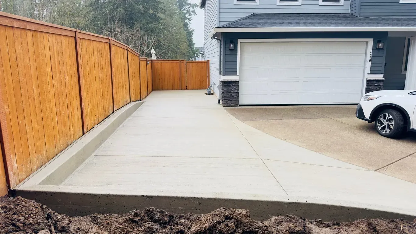 A newly poured concrete driveway extension sits next to a wooden fence beside a house with a white garage door.