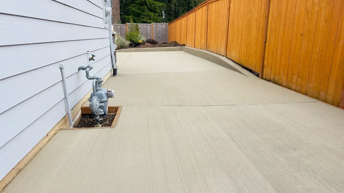 A newly poured concrete patio beside a white house, featuring a gas meter cutout and a wooden fence along the back.
