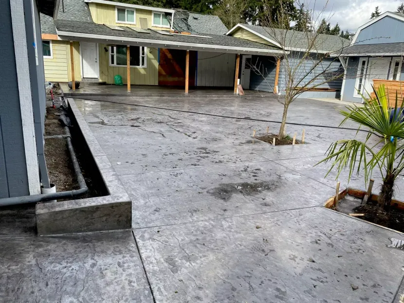 A wide view of a grey stamped concrete driveway or patio featuring a raised concrete planter box and a small palm tree.