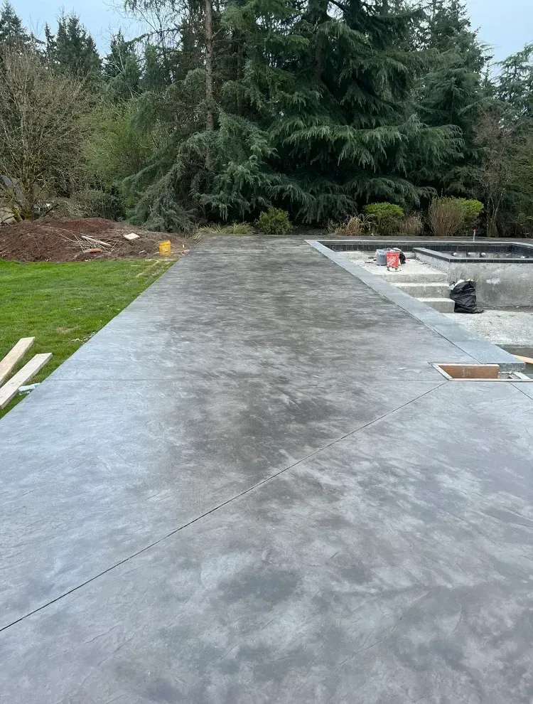 A freshly poured concrete patio stretches toward a backyard pool area with steps, surrounded by trees and a lawn.