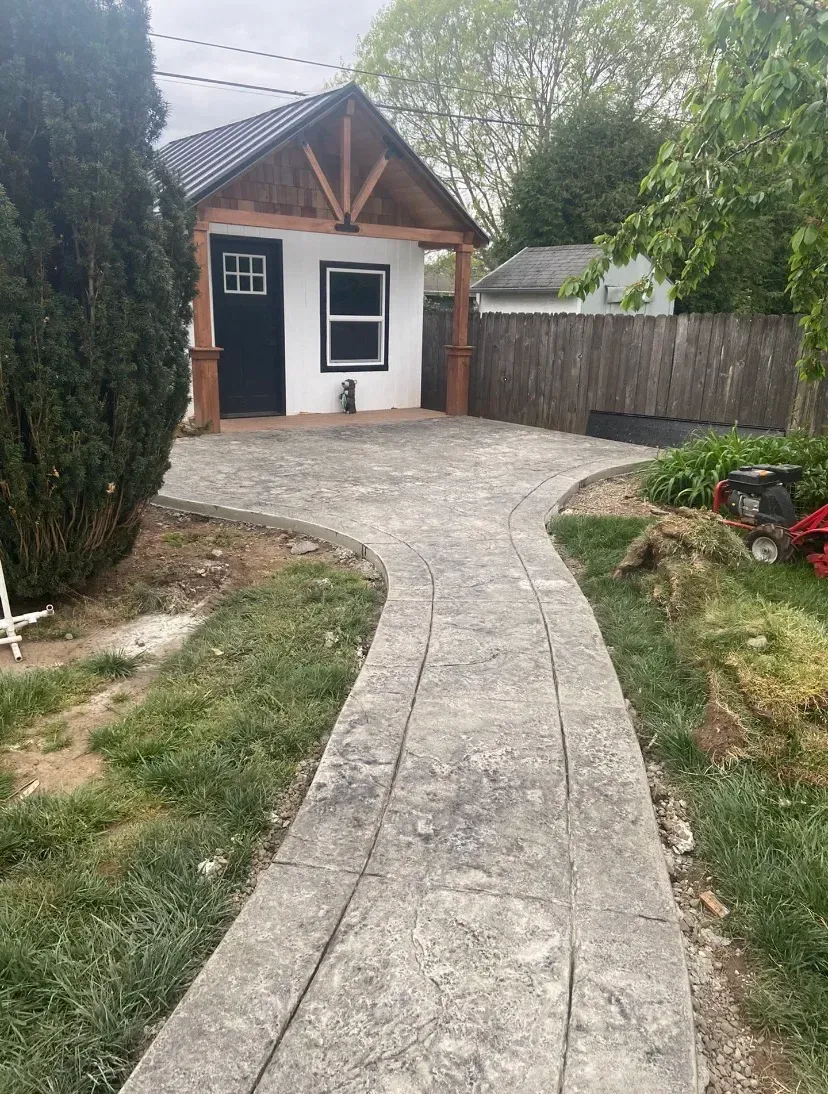 A stamped concrete walkway leads to a small, white building with a black door and a wooden front porch.