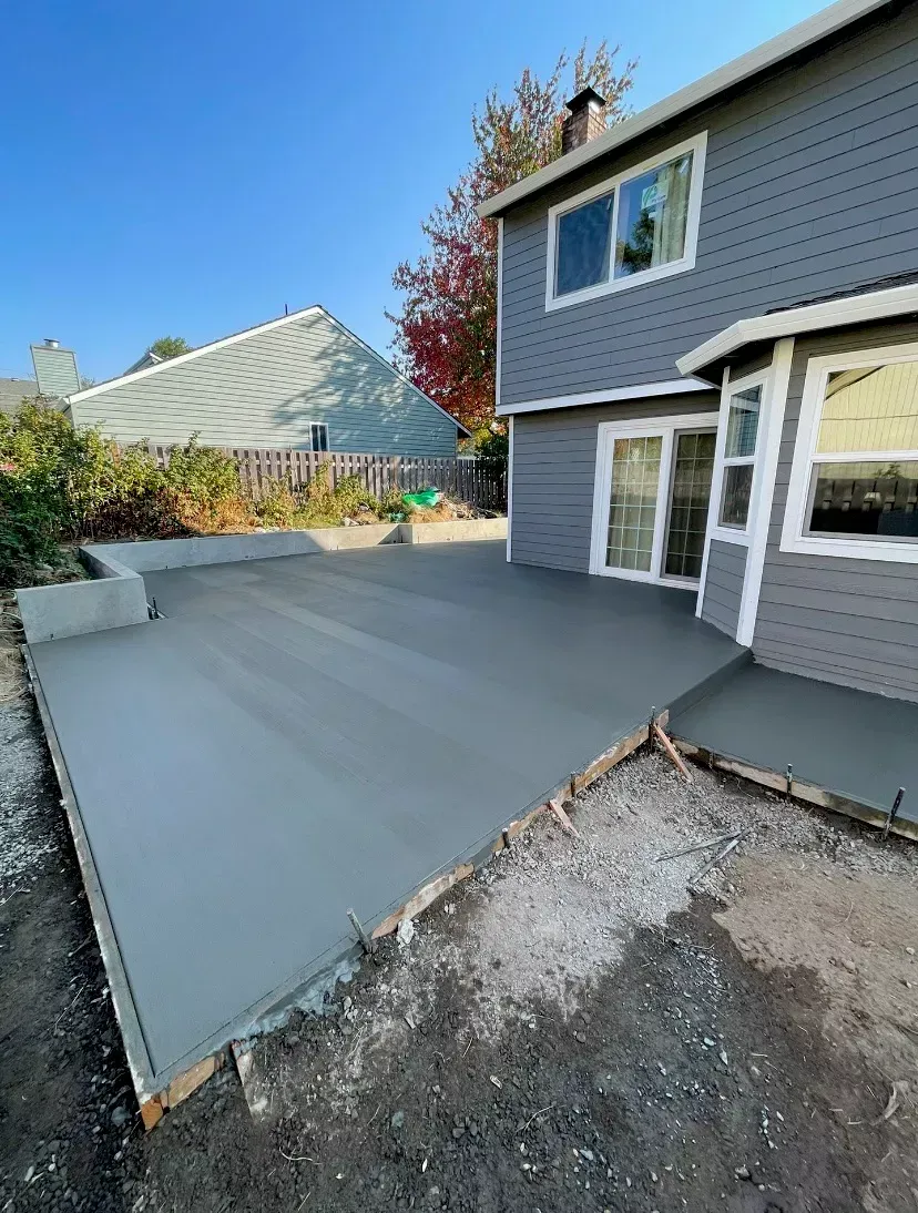 A newly poured gray concrete patio sits in the backyard of a blue house under a clear blue sky.