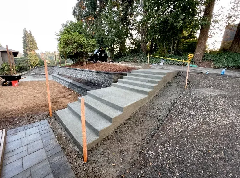 Newly poured concrete outdoor stairs set in a backyard landscape with unfinished ground and a paved area.