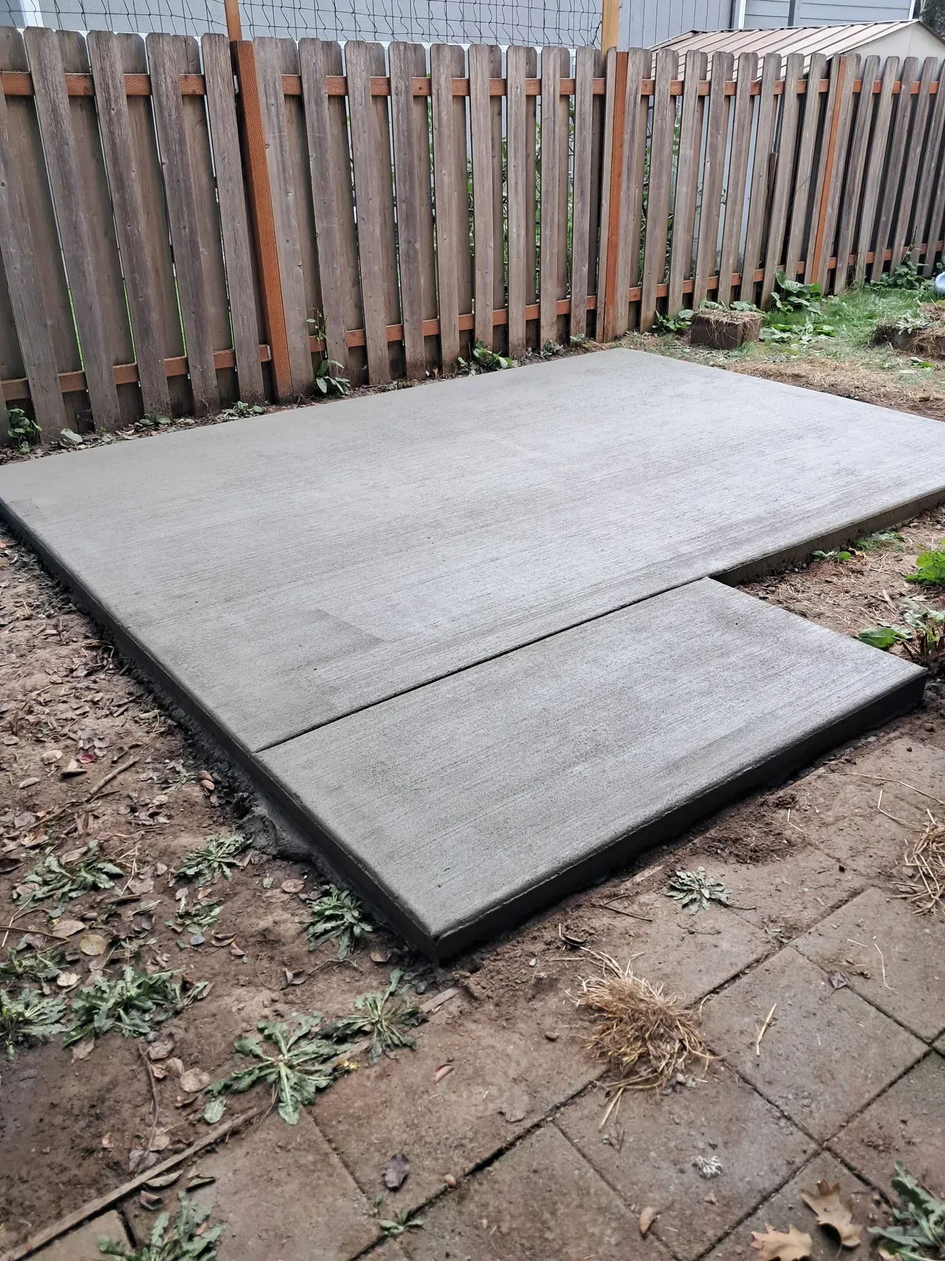 A newly poured, rectangular concrete patio slab situated in a backyard next to a wooden fence and paved stones.