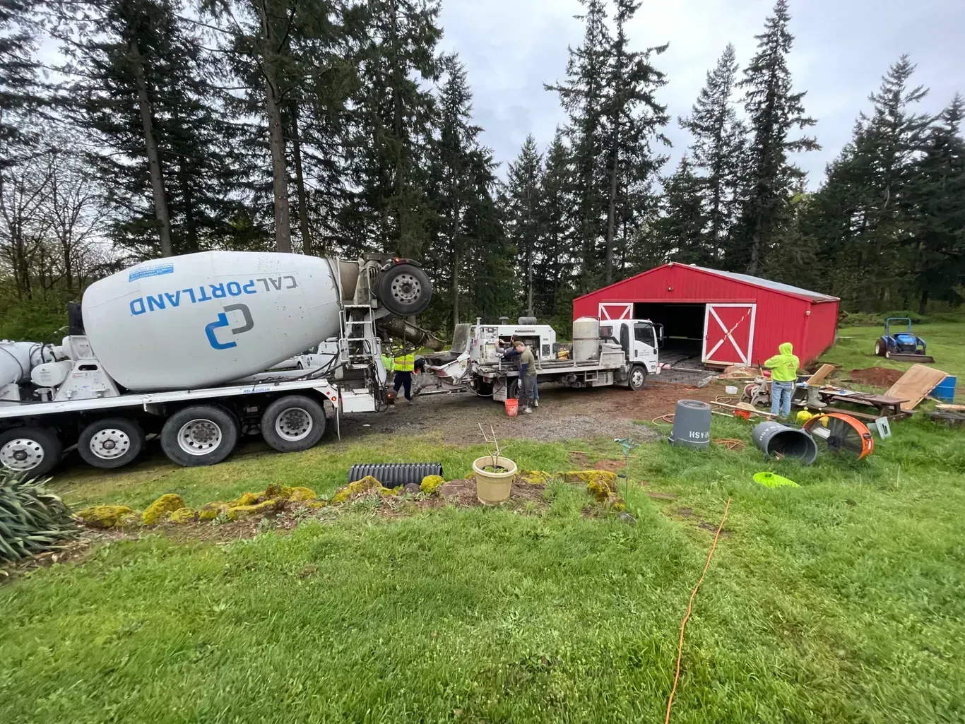 A cement truck and a construction vehicle parked near a red barn on a grassy property.