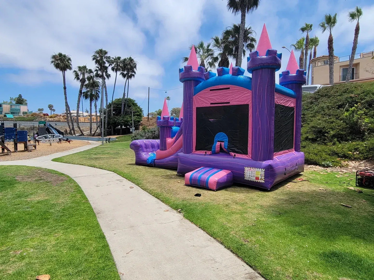 Purple inflatable bounce house on a grassy park lawn beside a paved path, with palm trees and a blue sky.