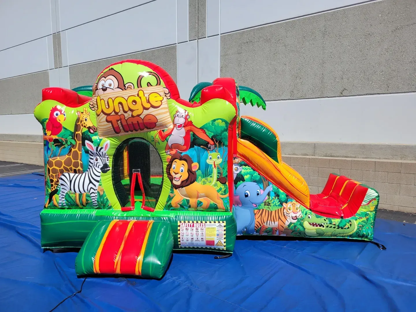 Jungle-themed inflatable bounce house with slide, bright green, red, and yellow, set up on a blue tarp outdoors