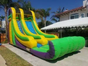 Green and yellow inflatable water slide in a sunny backyard next to a house and palm trees