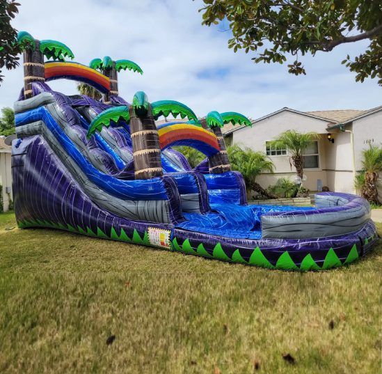 Large blue inflatable water slide in a grassy yard beside a house, with rainbow arches and palm tree accents