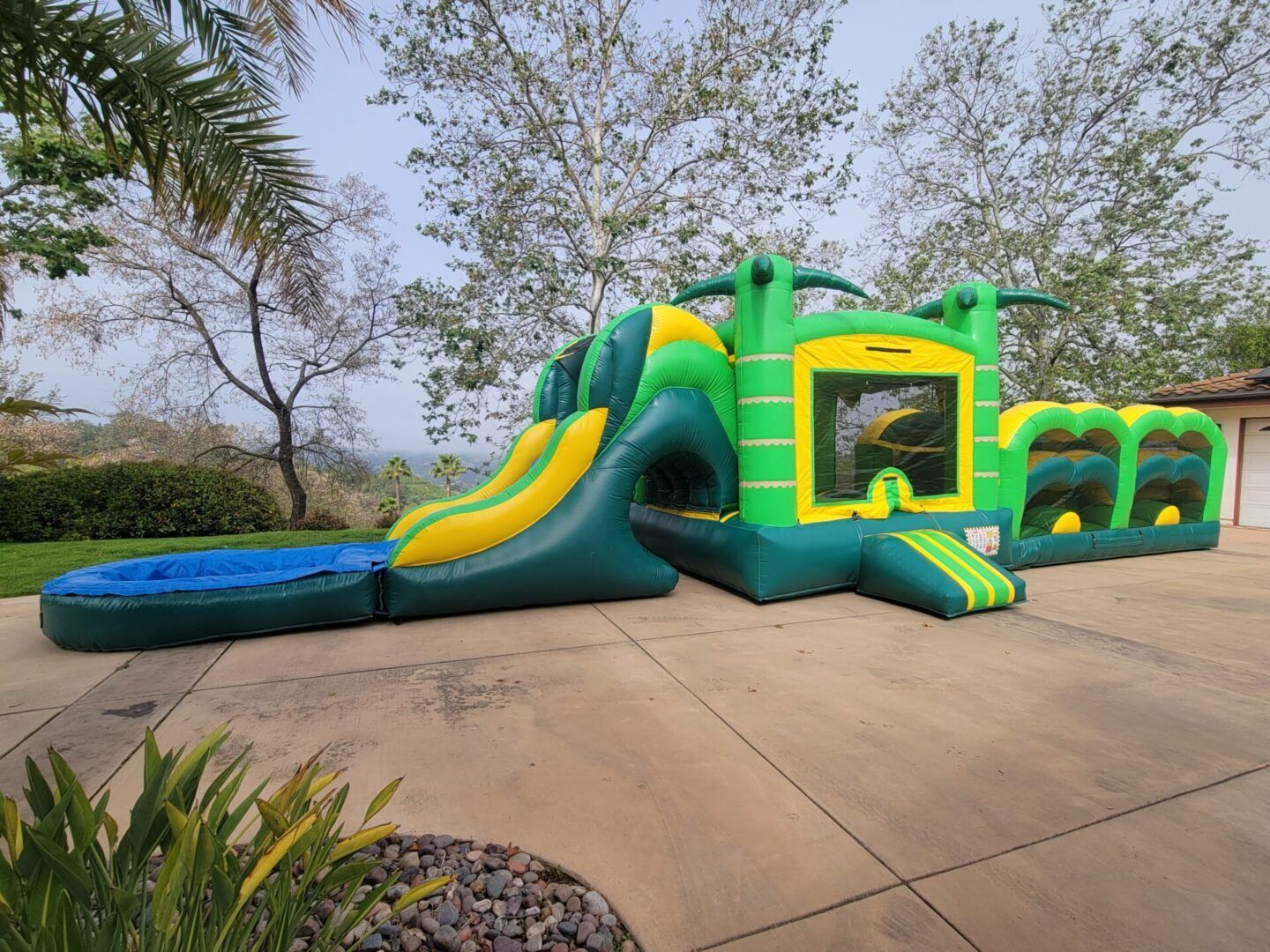 Green-and-yellow inflatable obstacle course with slide and pool in a paved backyard under trees