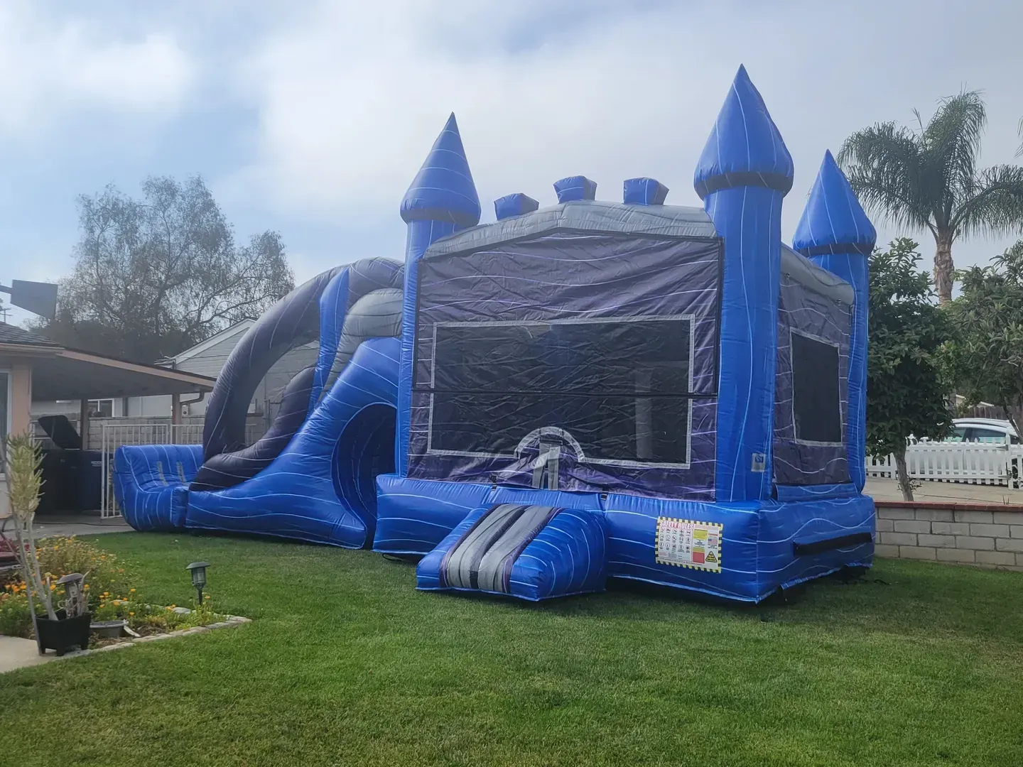 Blue inflatable bounce house with slide and towers set up on a grassy lawn outdoors.