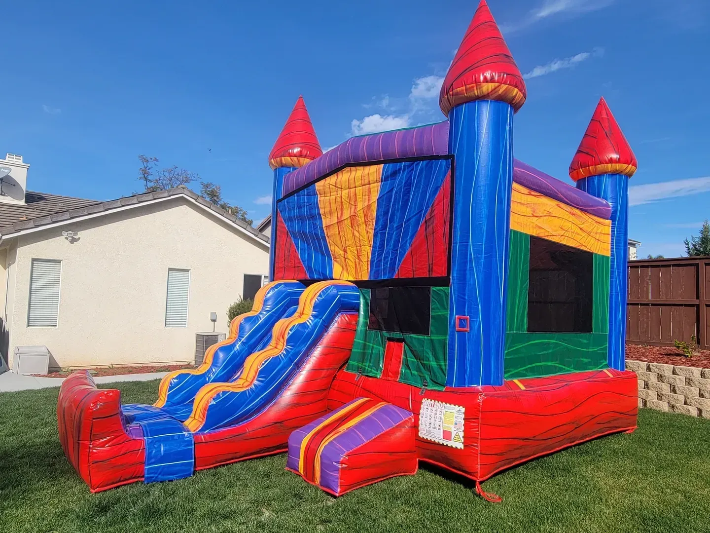 Colorful red and blue inflatable bounce house with slide on a grassy lawn beside a house
