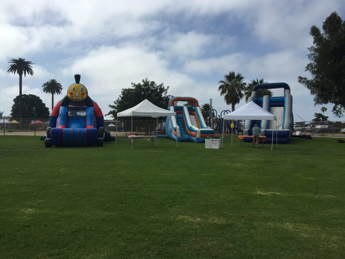 Inflatable bounce houses and slides on a grassy field under white tents on a cloudy day