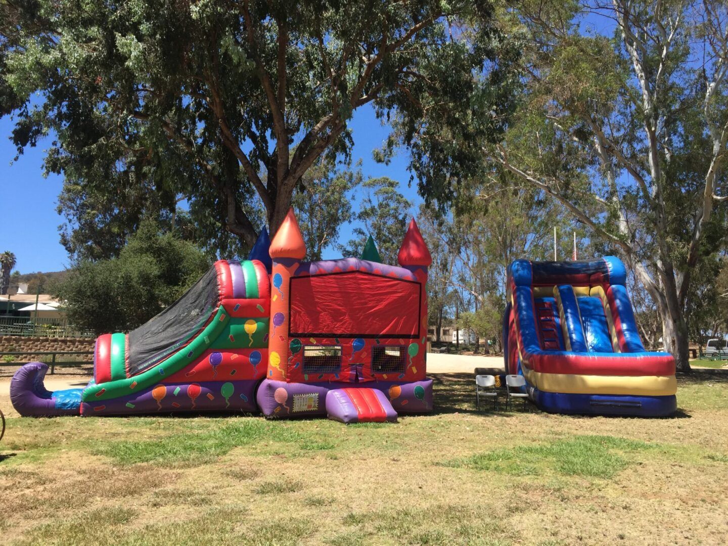 Two colorful inflatable bounce houses on a grassy field under trees