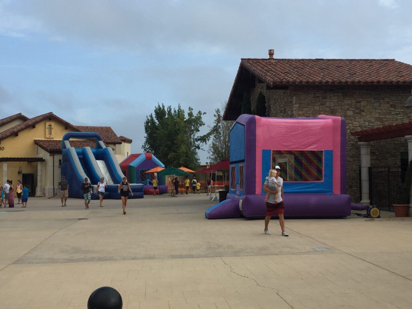 Inflatable bounce houses at an outdoor fair, with people playing in a courtyard.