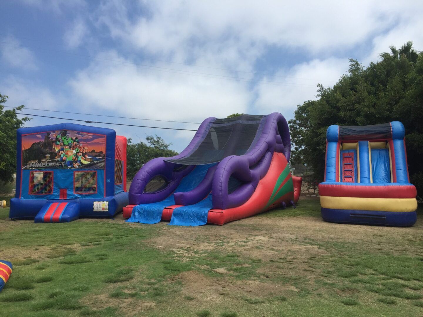 Three colorful inflatable bounce houses on grass under a cloudy sky at an outdoor event