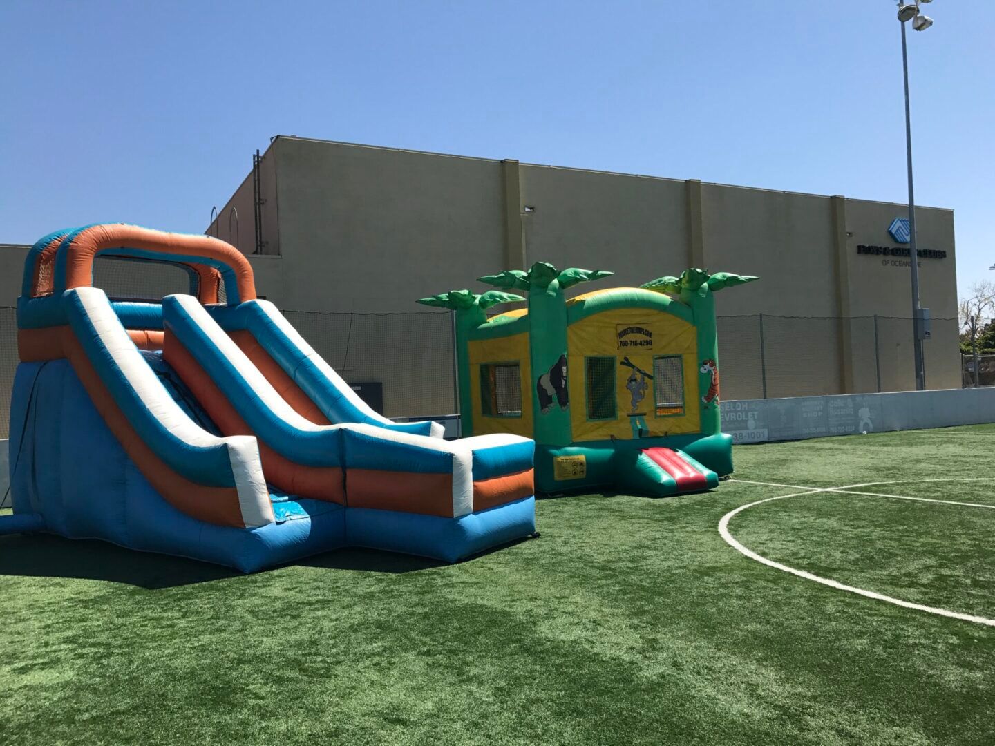 Two inflatable bounce houses on a grassy field under a clear blue sky with a large building in the background