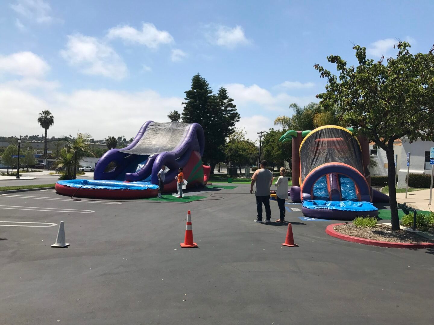 Two inflatable play structures in a parking lot, with cones and a few people under a sunny blue sky.