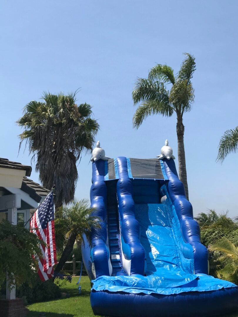 Blue inflatable water slide in a yard with palm trees and an American flag under a clear sky