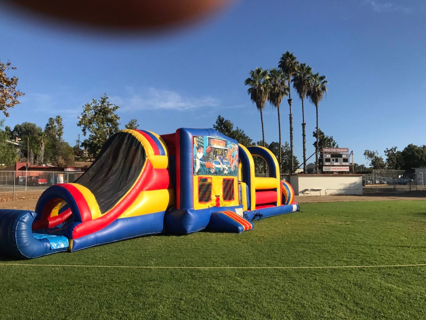 Colorful inflatable obstacle course on a grassy field under a blue sky with palm trees nearby