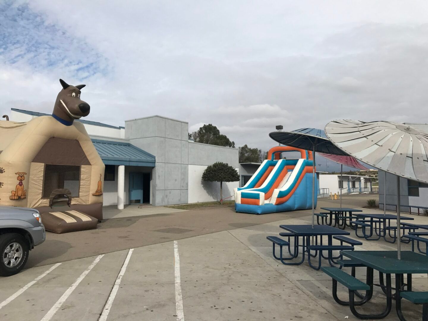 Outdoor play area with a giraffe-themed building, slide, and picnic tables under umbrellas