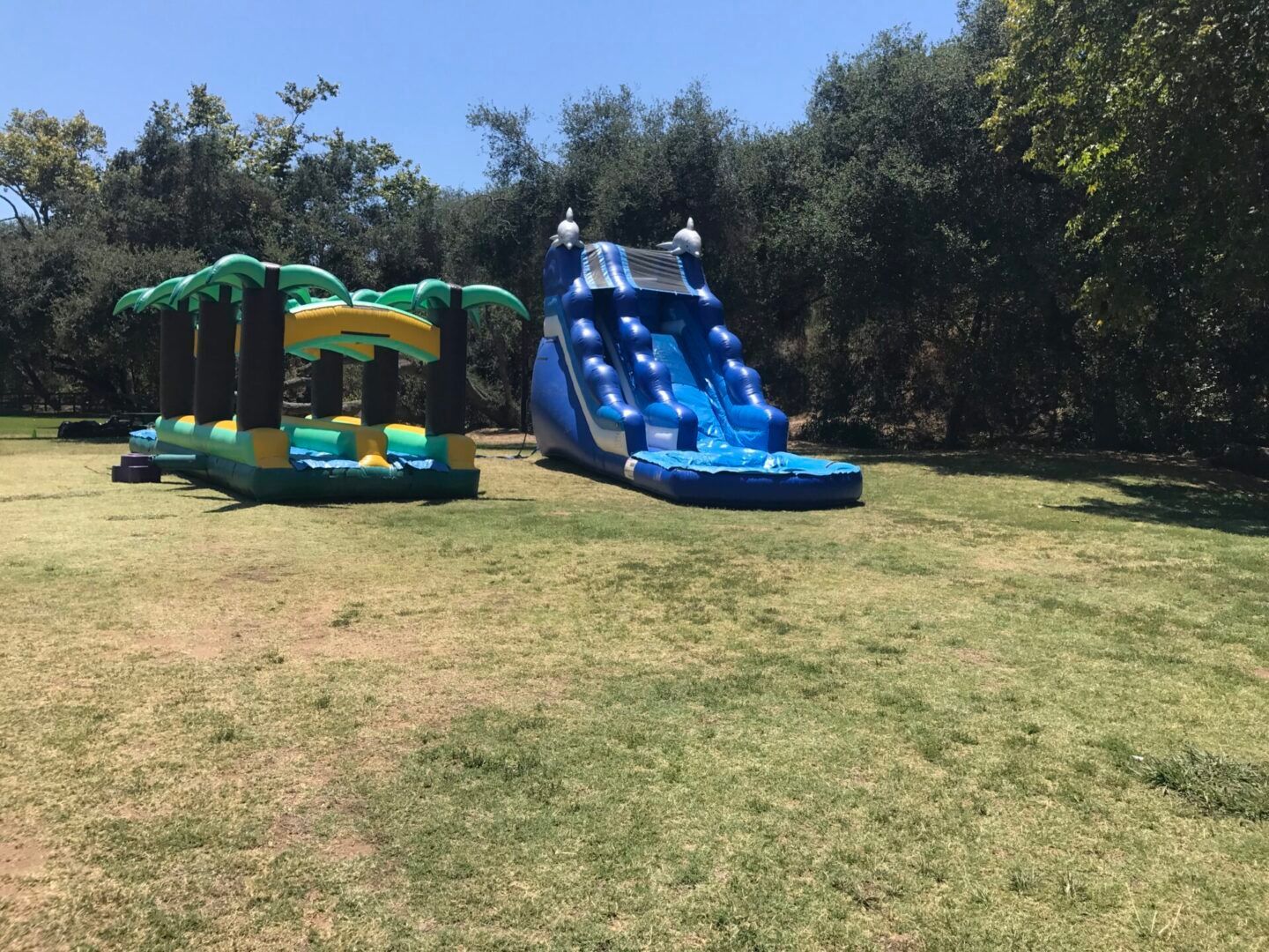 Two inflatable play structures on a grassy field, one green and yellow, one blue with slides.