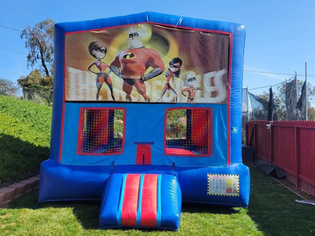 Blue inflatable bounce house with a superhero mural and red ramp in a grassy yard
