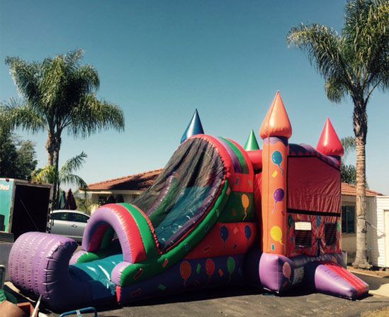 Colorful inflatable bounce house with slide in a sunny backyard, surrounded by palm trees