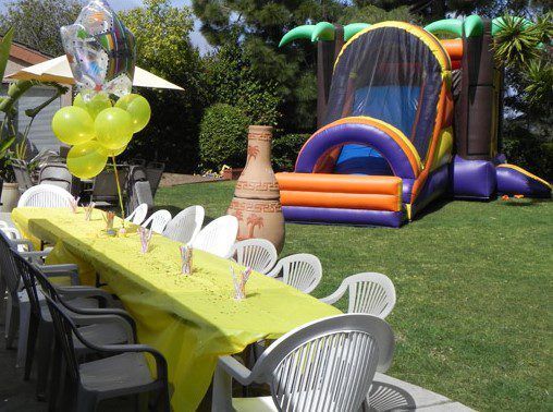 Outdoor party setup with long yellow table, chairs, balloons, and a colorful bounce house on a lawn