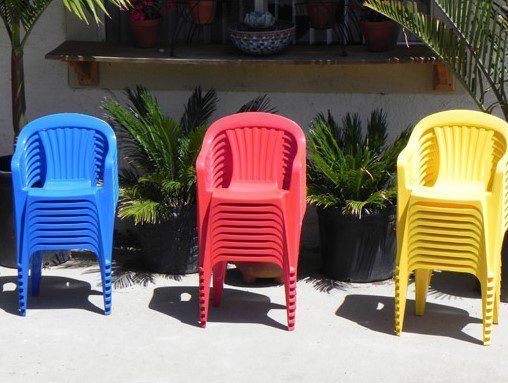 Three stacks of plastic chairs in blue, red, and yellow arranged outdoors in sunlight.