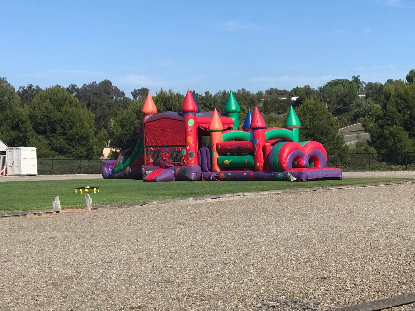 Colorful inflatable bounce house on a grassy field beside a gravel lot, with trees and blue sky behind.
