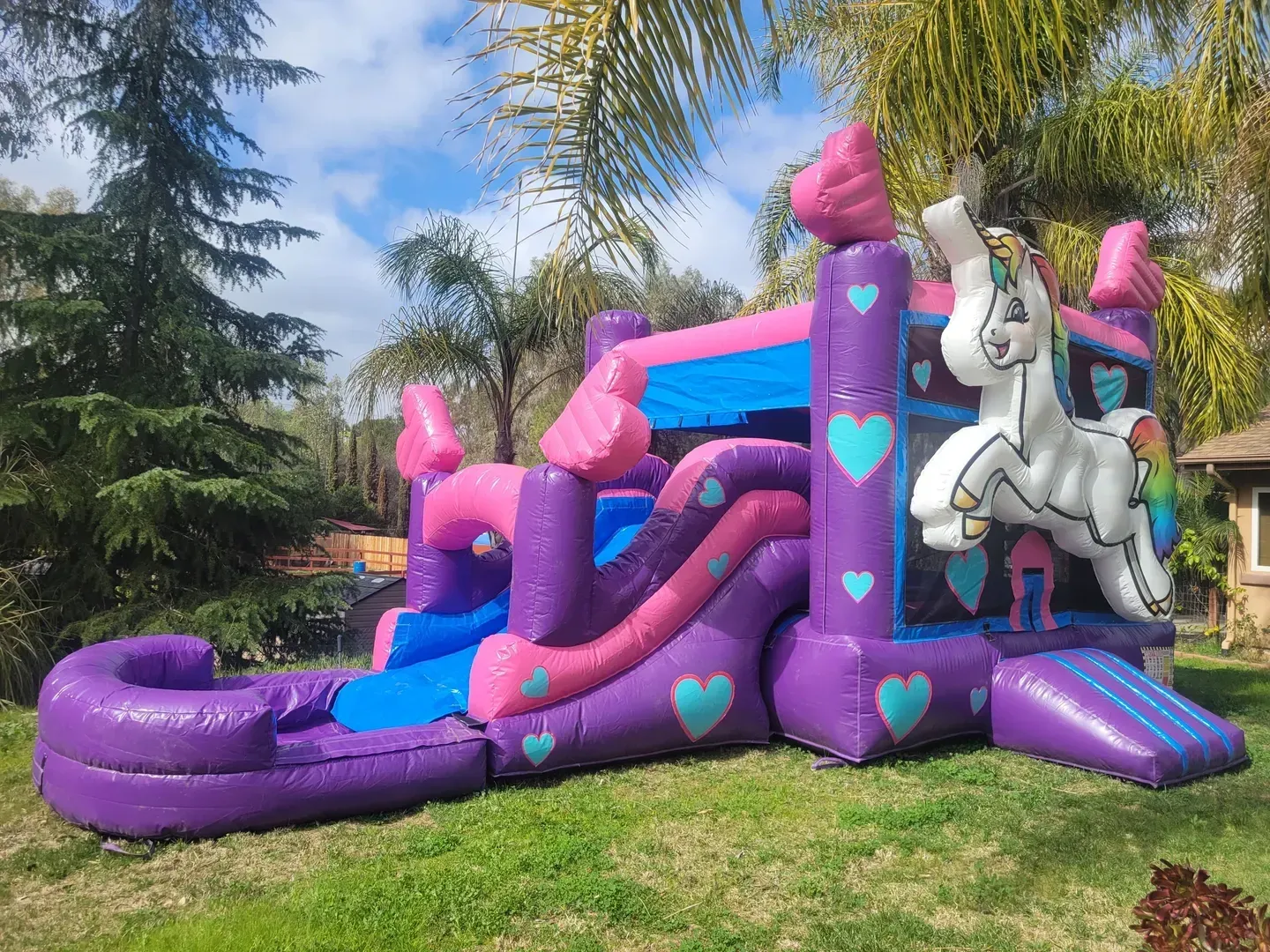 Purple-and-pink unicorn inflatable slide in a grassy yard, with heart decorations and palm trees nearby