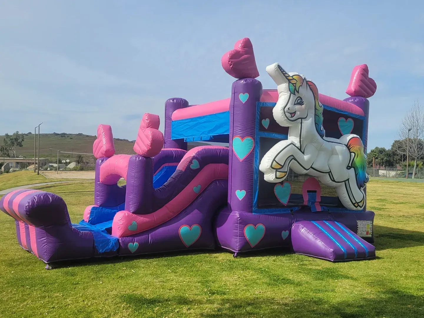 Pink and purple unicorn bounce house with slides on a grassy field under a blue sky