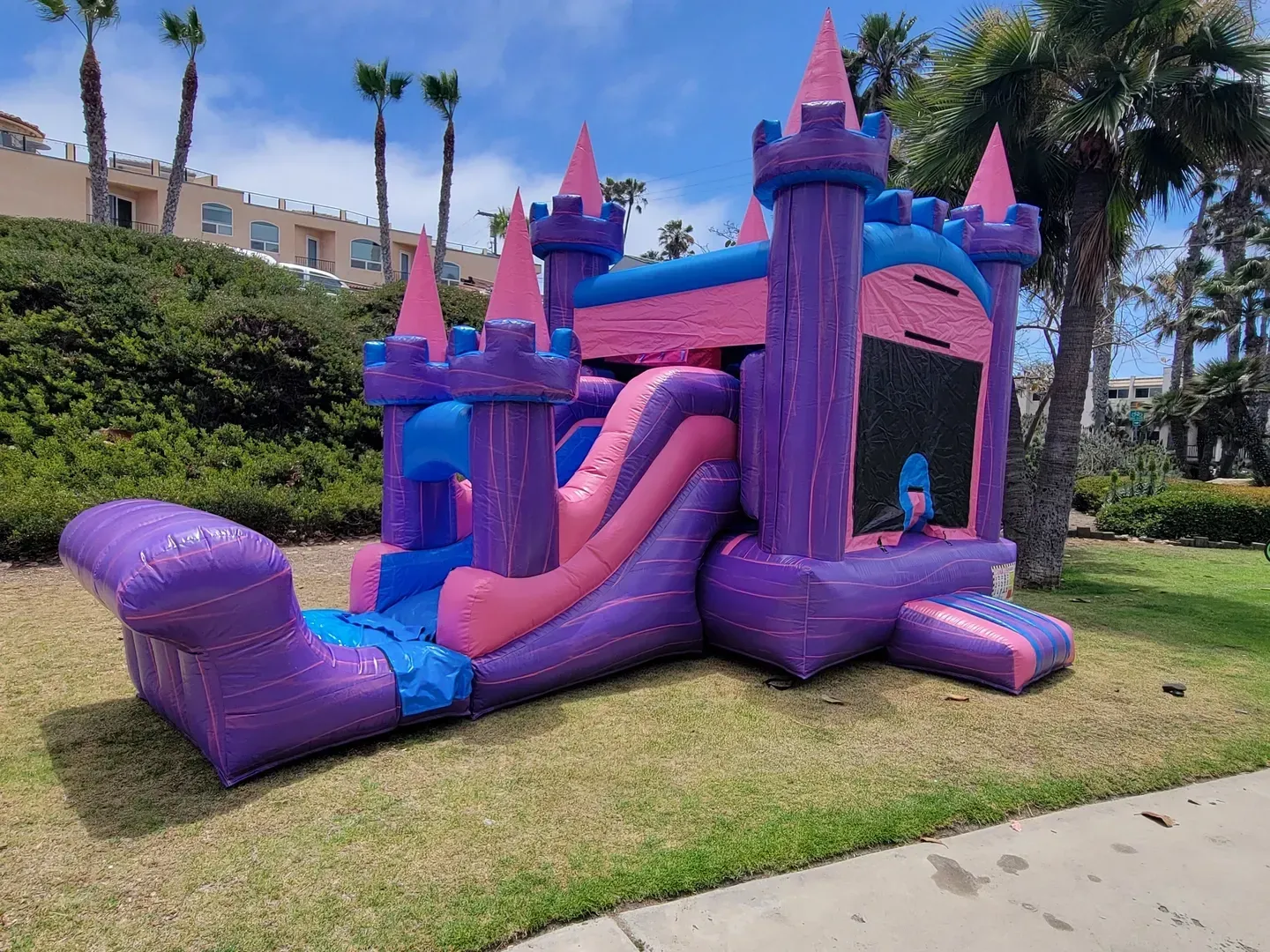 Purple inflatable castle-style bounce house with pink slide on a grassy lawn in a sunny park