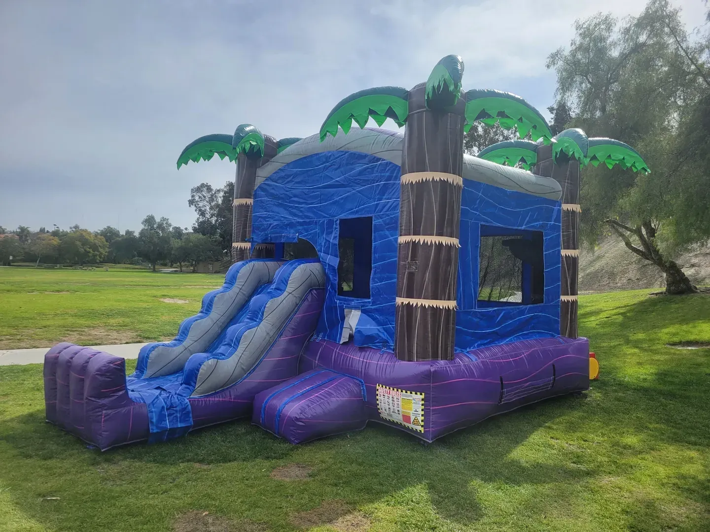 Blue and purple inflatable bounce house with slide and palm trees, set on grass outdoors