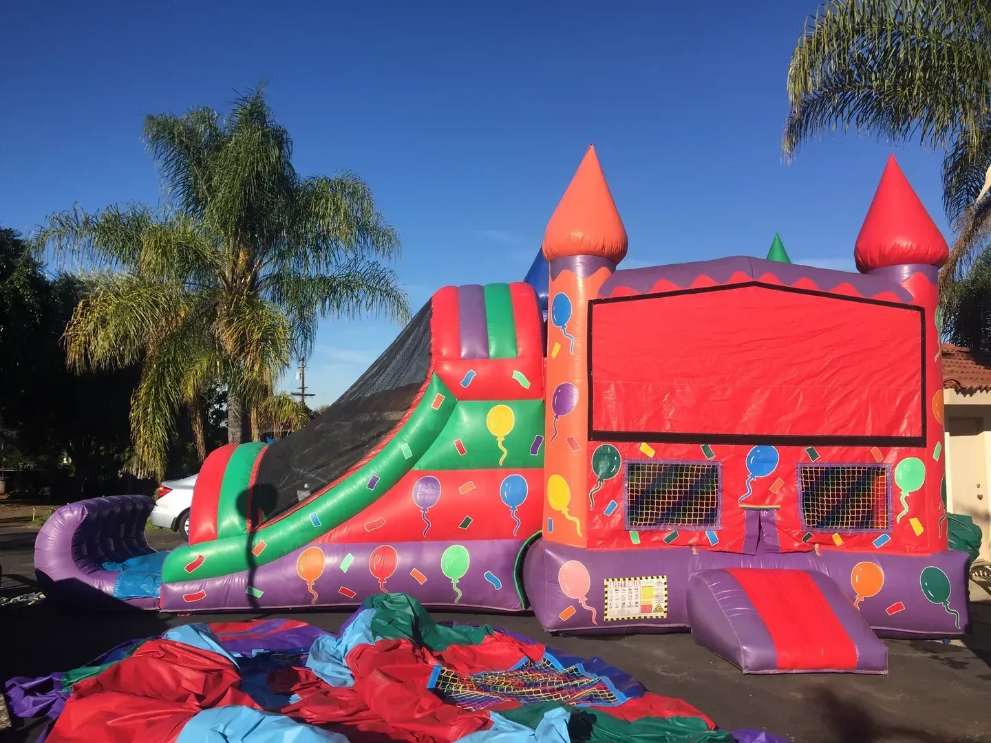 Colorful inflatable bounce house and slide in a sunny backyard, with palm trees and scattered toys nearby