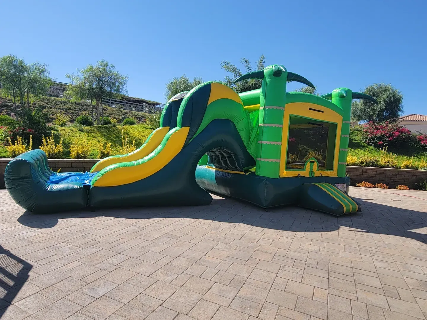 Green and yellow inflatable obstacle course slide on a paved patio with gardens in the background