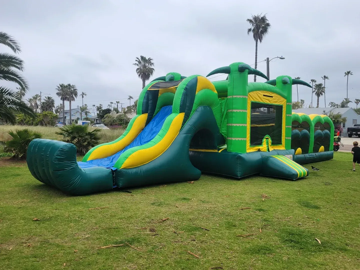 Green inflatable slide obstacle course on a grassy field with palm trees in the background