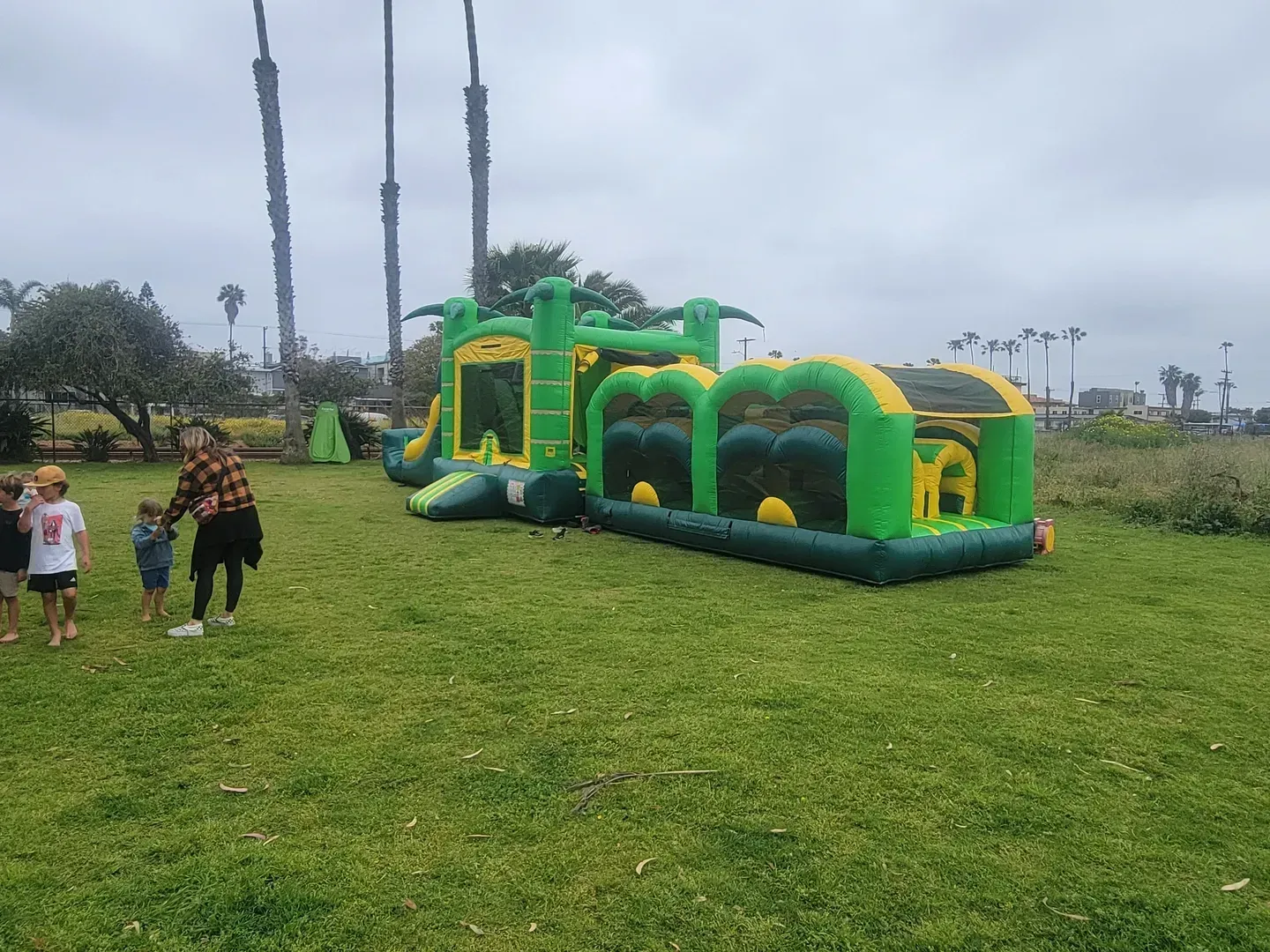 Green-and-yellow inflatable obstacle course on a grassy field with people nearby under a cloudy sky