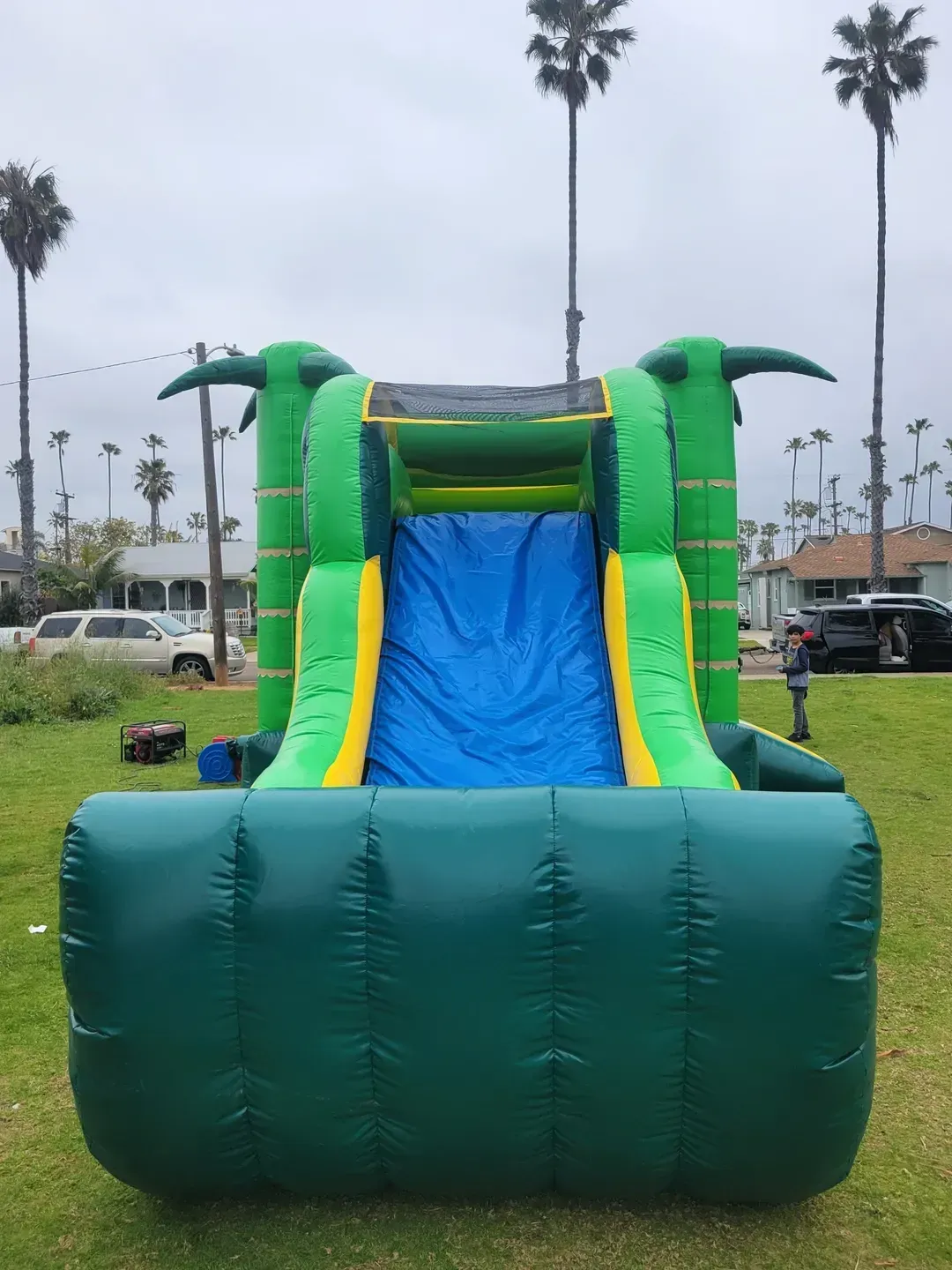 Green inflatable slide bounce house on grass, with blue slide and palm trees in the background