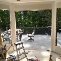 Open doorway to a sunny deck with chairs, table, railing, and trees beyond