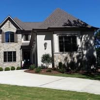 Large two-story brick house with gray roof, white driveway, and green lawn in sunlight