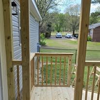 Wooden porch with railing overlooking a grassy yard and two parked cars beyond.